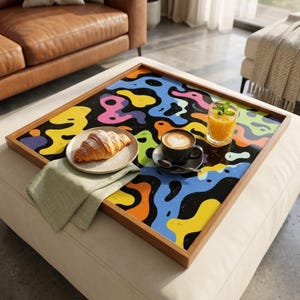 May include: A wooden serving tray with a vibrant abstract pattern in yellow, blue, orange, and purple on a black base. It displays a croissant, coffee, and juice. The tray rests on a white ottoman.