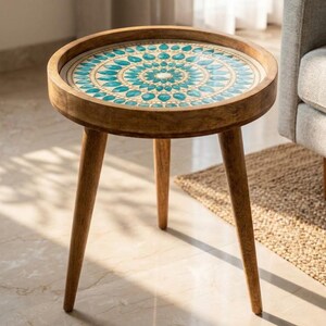 May include: A round wooden side table featuring three tapered legs and a circular tabletop. The surface of the table is inlaid with a decorative pattern of teal and white teardrop-shaped tiles arranged in a concentric mandala-like design.