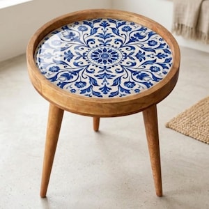 Op de afbeelding: Ronde houten bijzettafel met een blauw-wit bloemenpatroon op het tafelblad. De tafel heeft drie schuine poten en een opstaande rand. Het hout heeft een natuurlijke afwerking. De diameter is ongeveer 40 cm.