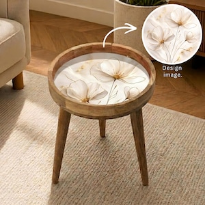 May include: A round wooden side table with three tapered legs and a flat top surface featuring a printed graphic of white flowers with thin brown stems and gold centers. The design image is shown in a circular inset with the text Design image.