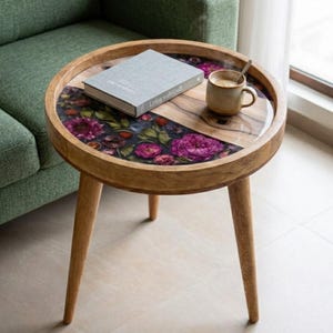 May include: A round wooden side table with three tapered legs. The tabletop features a split design with one half showing natural wood grain and the other half inlaid with a dark resin floral pattern. A grey book and a ceramic mug sit on top.