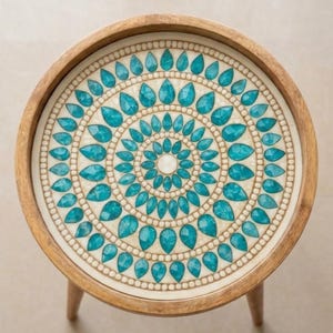 May include: A round wooden side table with a light brown frame and a circular tabletop featuring a decorative inlay. The inlay consists of concentric rings of teal, teardrop-shaped stones set against a cream-coloured background with small beads.