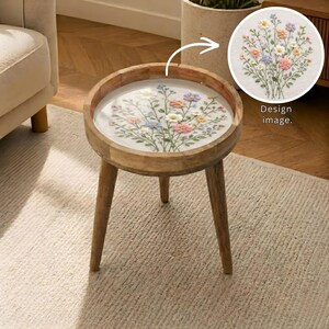Puede incluir: Una mesa auxiliar redonda de madera con un diseño floral bordado en la encimera. La mesa tiene tres patas anguladas y un borde elevado. El diseño presenta flores coloridas, incluyendo rosa, amarillo y azul, sobre un fondo blanco. El texto "Design image" es visible.