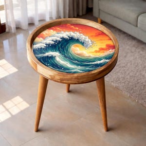 May include: A round side table with a wooden frame and a painted tabletop featuring a dynamic ocean wave scene. The wave is rendered in blue and white, set against a sunset. The table has three wooden legs.