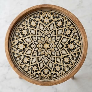 Pu&ograve; includere: Un tavolino rotondo in legno con un piano intarsiato caratterizzato da un motivo geometrico a stella. Il design &egrave; composto da segmenti alternati color crema, nero e marrone chiaro disposti in simmetria radiale, incorniciati da un bordo in legno naturale.