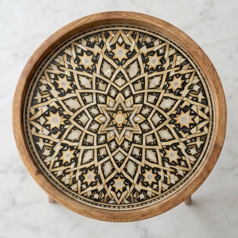 Pu&ograve; includere: Un tavolino rotondo in legno con un piano intarsiato caratterizzato da un motivo geometrico a stella. Il design &egrave; composto da segmenti alternati color crema, nero e marrone chiaro disposti in simmetria radiale, incorniciati da un bordo in legno naturale.