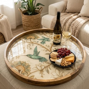 May include: A round wooden serving tray with a hummingbird and floral design. The tray holds a bottle of wine, a glass of wine, a plate of snacks, and is set on a neutral-colored ottoman. The tray has a clear, glossy surface.