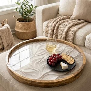 Peut inclure: Un plateau de service rond en bois avec une surface transparente à motifs ondulés. Il contient un verre de vin blanc, une petite assiette noire avec des raisins, un cracker et du fromage. Le plateau est sur un pouf de couleur neutre.
