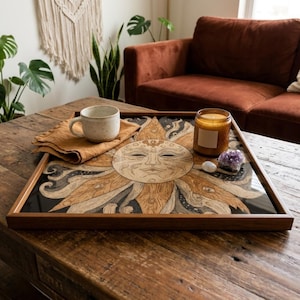 May include: A wooden tray with a sun design, a mug, a napkin, a candle, and crystals on a wooden coffee table. The tray has a dark brown frame and a detailed sun illustration in shades of brown and cream. The mug is speckled, and the candle is in a glass jar.