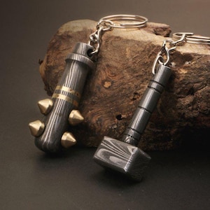 Könnte beinhalten: Zwei Schlüsselanhänger mit filigranen Metallarbeiten. Ein Schlüsselanhänger ist ein Stachelschläger, der andere ein Hammer. Beide sind aus dunklem Metall mit einem wirbelnden Muster.