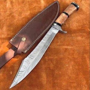 May include: A Damascus steel knife with a serrated edge and a wooden handle, resting on a brown surface. The knife is accompanied by a brown leather sheath. The handle has black bands. The knife is a hunting or survival tool.