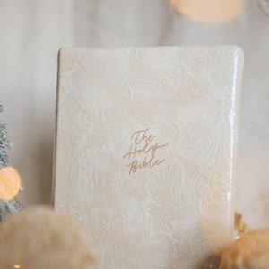 Op de afbeelding: Een crèmekleurige Bijbel met een bloemreliëf. De titel "The Holy Bible" is in gouden letters geschreven. Het boek staat rechtop, met zachte, vage decoraties op de voorgrond.