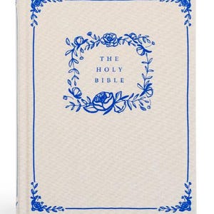 May include: A hardcover book with a light beige fabric cover featuring a vibrant blue floral border and a central wreath design. The text THE HOLY BIBLE is printed in blue in the center of the wreath. The cover has a clean, simple aesthetic.