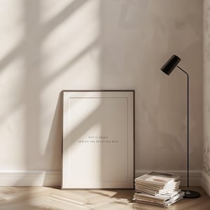 May include: A framed print with the text "hell is empty and all the devils are here" in a minimalist style. The print is leaning against a wall, next to a stack of books and a black floor lamp. The room has a neutral colour palette.