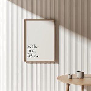 May include: A framed print with the text "yeah, fine, fck it" in a minimalist style. The print is in a light brown frame and hangs on a white, textured wall. A small wooden table and two small gray cups are in the foreground.