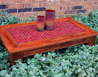 May include: A wooden coffee table with a red tile top. The table is sitting on a bed of green plants. Two red glass candle holders are on the table.