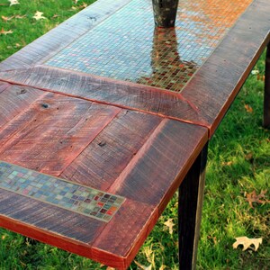 K&ouml;nnte beinhalten: Ein rustikaler Holztisch mit einer Mosaikplatte. Der Tisch hat eine dunkle Holzoptik und ist aus wiederverwendetem Holz gefertigt. Die Mosaiksteine sind in Braun-, Gr&uuml;n- und Rott&ouml;nen gehalten. Der Tisch steht auf einem gr&uuml;nen Rasen.