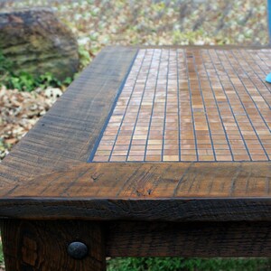 May include: A square wooden table with a mosaic tile top. The tiles are a light brown colour and are arranged in a grid pattern. The table has a dark brown wood frame and legs.