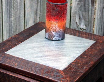 May include: A small wooden end table with a metal top. The table has a dark brown stain and a distressed finish. A red, orange, and white glass candle holder sits on the table.