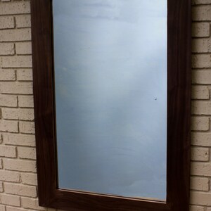 May include: A dark brown wooden frame with a mirror reflecting a cloudy sky. The frame is mounted on a brick wall.