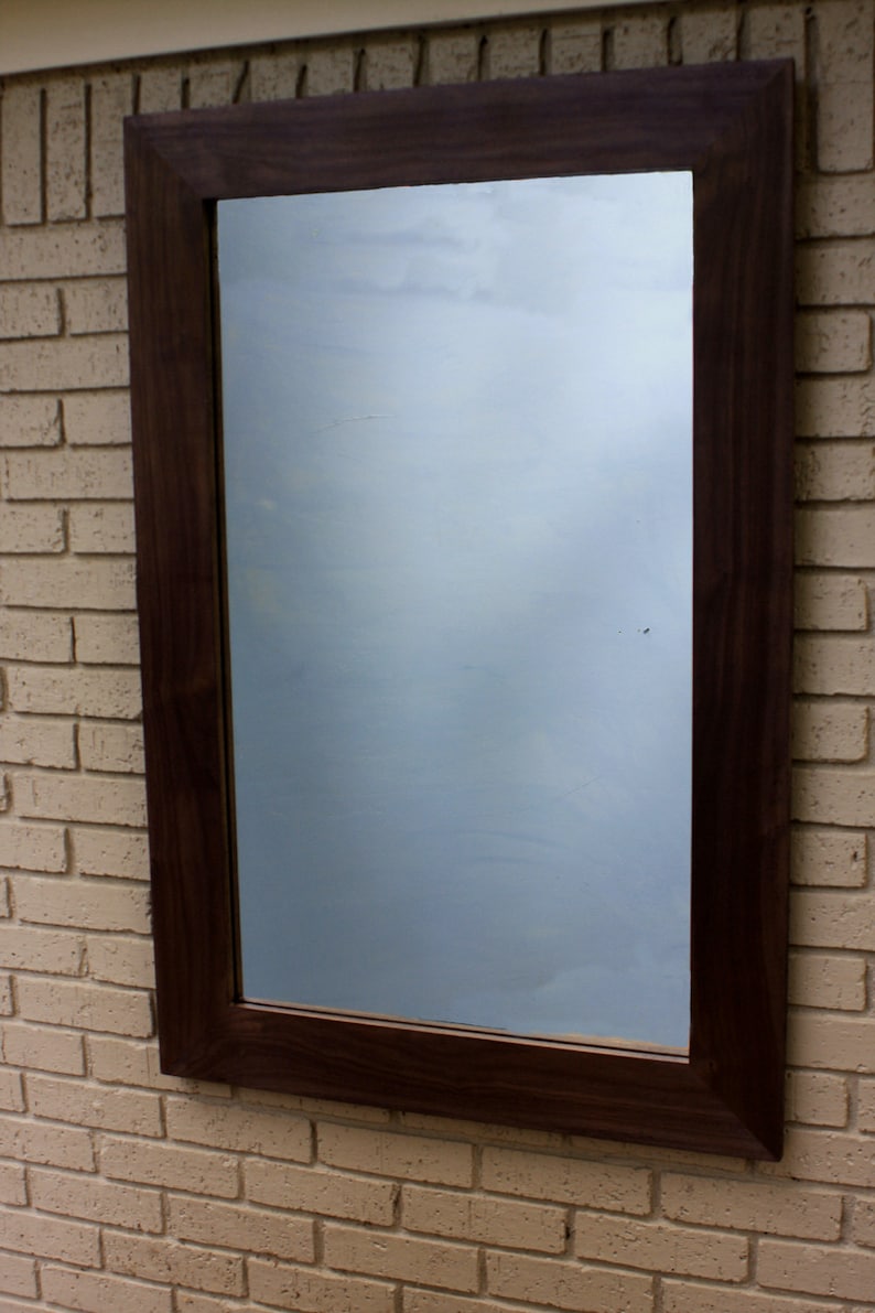 May include: A dark brown wooden frame with a mirror reflecting a cloudy sky. The frame is mounted on a brick wall.