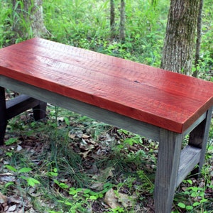 May include: A rustic wooden bench with a red stained top and gray stained legs. The bench is sitting on a bed of leaves and grass.