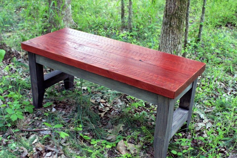 May include: A rustic wooden bench with a red stained top and gray stained legs. The bench is sitting on a bed of leaves and grass.