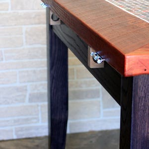 K&ouml;nnte beinhalten: Ein Holztisch mit dunkel gebeizter Oberfl&auml;che und einer Mosaik-Tischplatte. Der Tisch hat ein schlichtes Design mit geraden Beinen und einer rechteckigen Platte. Die Mosaiksteine sind in einem Gittermuster angeordnet und weisen eine Vielzahl von Farben auf, darunter Rot, Braun und Wei&szlig;.