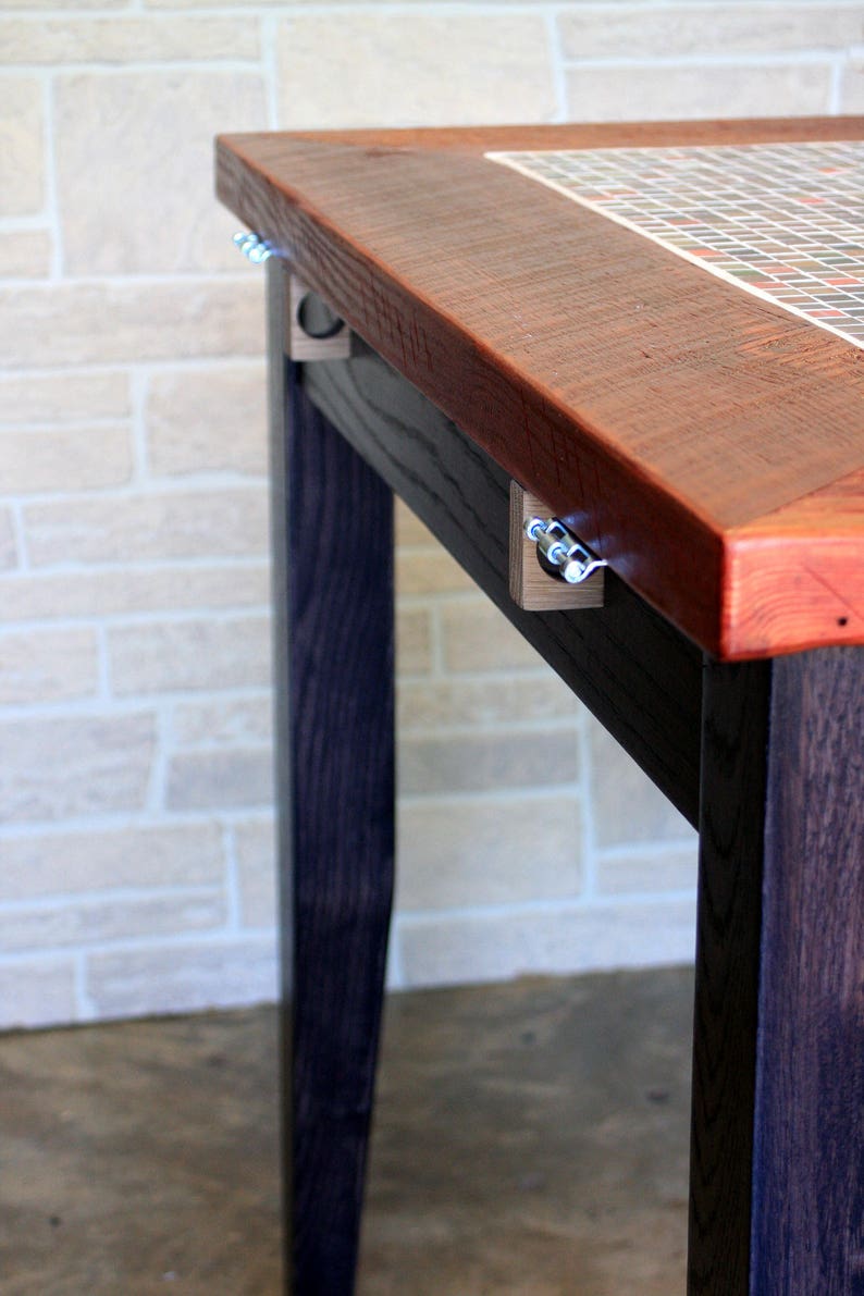 K&ouml;nnte beinhalten: Ein Holztisch mit dunkel gebeizter Oberfl&auml;che und einer Mosaik-Tischplatte. Der Tisch hat ein schlichtes Design mit geraden Beinen und einer rechteckigen Platte. Die Mosaiksteine sind in einem Gittermuster angeordnet und weisen eine Vielzahl von Farben auf, darunter Rot, Braun und Wei&szlig;.