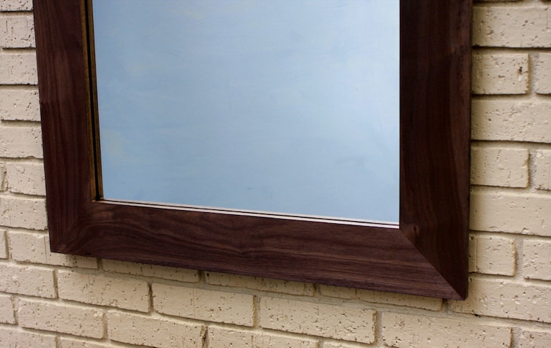 May include: A dark brown wooden frame with a mirror inside. The frame is mounted on a white brick wall.