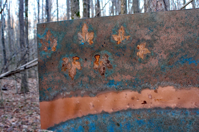 May include: A metal sheet with a blue and brown patina. The sheet has a horizontal line of copper running across the middle. There are several leaf shapes pressed into the metal.