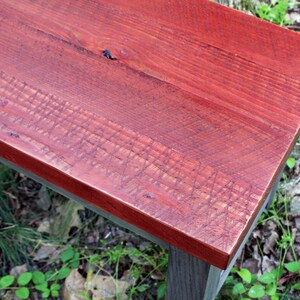 May include: A close-up of a wooden bench with a red stained top. The bench has a dark gray base and is made of reclaimed wood.