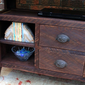 May include: A dark brown wooden entertainment center with two drawers and shelves. The drawers have ornate metal handles. The shelves are empty except for a bowl of blue stones and a stack of books.