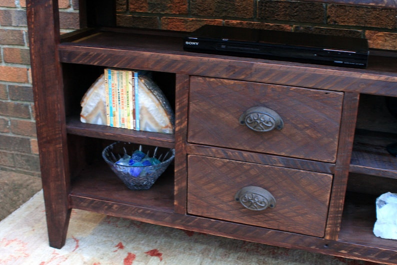 May include: A dark brown wooden entertainment center with two drawers and shelves. The drawers have ornate metal handles. The shelves are empty except for a bowl of blue stones and a stack of books.