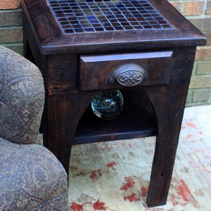 May include: A dark wood end table with a drawer and a shelf. The top of the table is covered in a mosaic of blue and purple tiles. The table has a decorative metal knob on the drawer.