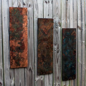 May include: Three rectangular metal wall art pieces with a rustic, weathered look. The pieces are in shades of brown, green, and blue, with a mottled, textured surface.