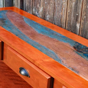 May include: A wooden console table with two drawers and a copper-colored top. The copper top has a blue and brown patina. The drawers have dark metal handles. The table is made of reddish-orange wood.