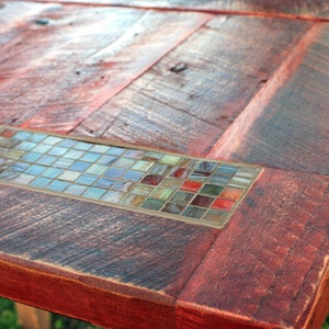 K&ouml;nnte beinhalten: Eine rustikale Holztischplatte mit einem Mosaik-Einlegeband. Die Tischplatte ist aus wiederverwendetem Holz und hat eine r&ouml;tlich-braune Farbe. Das Mosaik-Einlegeband besteht aus kleinen, bunten Glasfliesen.