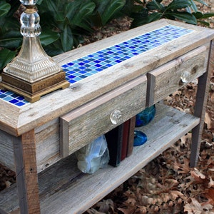 May include: A rustic wooden console table with two drawers and a lower shelf. The top of the table is decorated with a mosaic of blue and green tiles. The table is sitting on a bed of brown leaves.