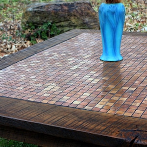May include: A wooden table with a copper mosaic top. The table has a dark brown wood frame and a light brown mosaic top. A blue vase is sitting on the table.