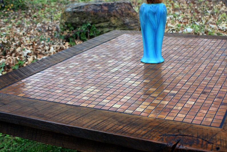 May include: A wooden table with a copper mosaic top. The table has a dark brown wood frame and a light brown mosaic top. A blue vase is sitting on the table.
