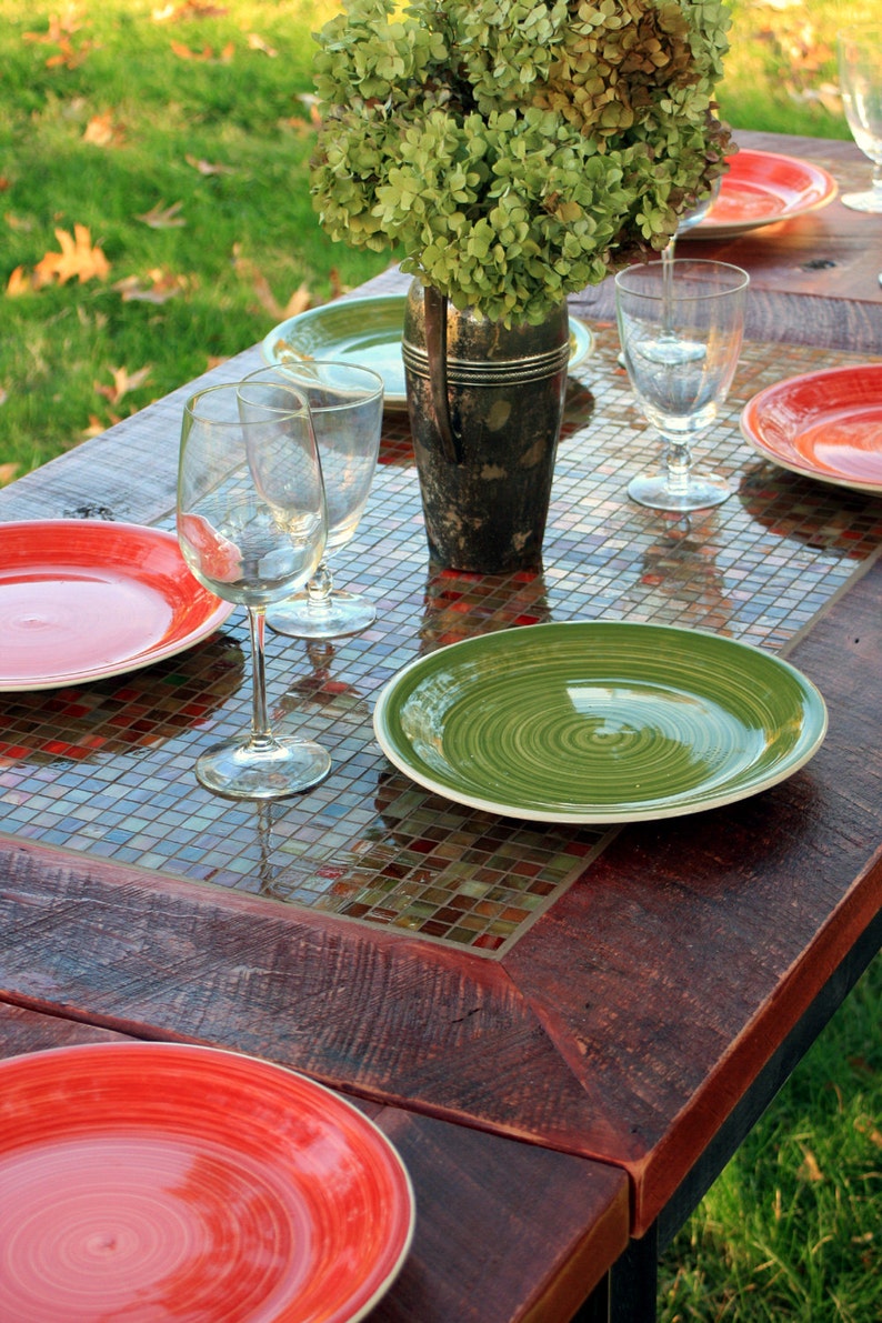 K&ouml;nnte beinhalten: Ein rustikaler Holztisch mit einer Mosaik-Tischplatte. Der Tisch ist mit roten und gr&uuml;nen Tellern, Weingl&auml;sern und einer Vase mit gr&uuml;nen Blumen gedeckt.
