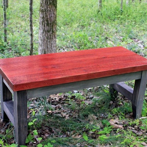 May include: A rustic wooden bench with a red stained top and gray stained legs. The bench is sitting in a grassy area with trees in the background.