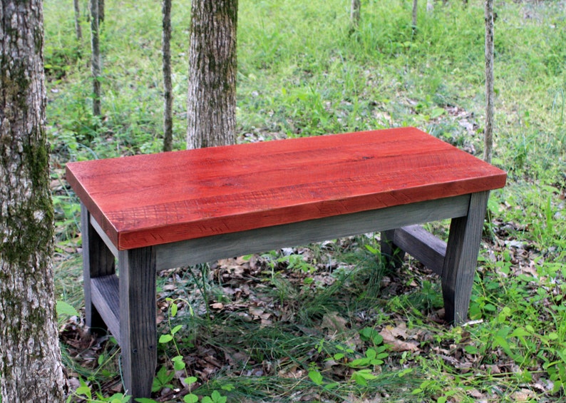 May include: A rustic wooden bench with a red stained top and gray stained legs. The bench is sitting in a grassy area with trees in the background.