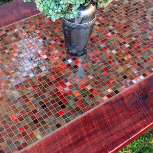 K&ouml;nnte beinhalten: Ein Holztisch mit einer Mosaik-Tischplatte. Die Fliesen sind klein und in den Farben Rot, Orange, Gr&uuml;n und Braun gehalten. Eine silberne Vase mit einem Strau&szlig; wei&szlig;er Blumen steht auf dem Tisch.