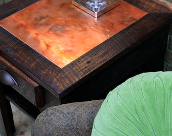 May include: A dark wood end table with a copper top and a crystal lamp on top. The table has a drawer and is sitting on a rug. A green velvet pillow is on a chair in the background.