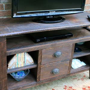May include: A dark brown wooden TV stand with two drawers and open shelves. The stand has a distressed finish and features decorative metal drawer pulls. A flat screen TV and a DVD player are on top of the stand.