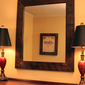 May include: A dark brown wooden framed mirror with a beveled edge, hanging on a yellow wall above a dark wood dresser. Two black lamps with red bases are on either side of the mirror. The mirror reflects a yellow wall with a framed print of a vintage wine label.