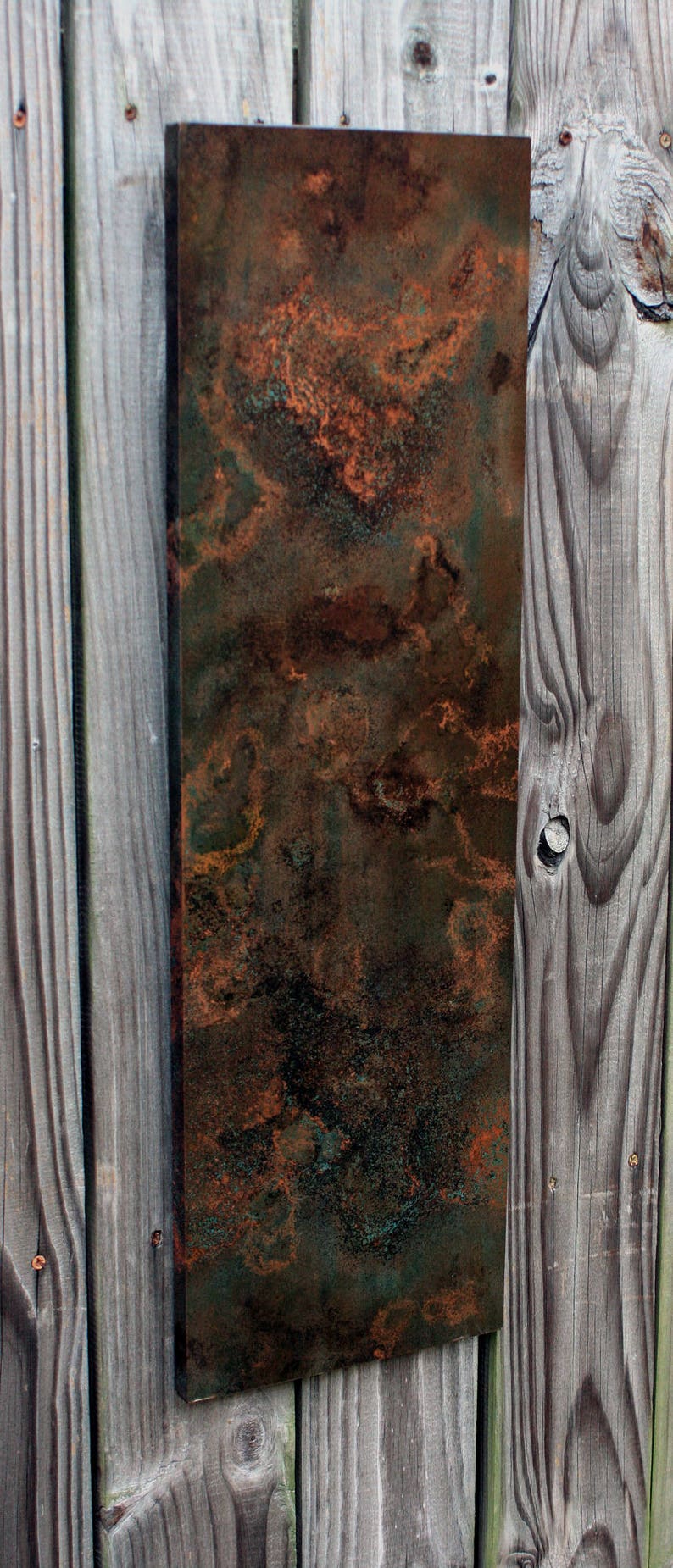 May include: A rectangular metal panel with a mottled, rusty, and green patina. The panel is mounted on a wooden wall.
