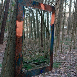 May include: A rectangular mirror with a copper frame, showing a reflection of a forest. The frame has a blue and green patina, and the mirror is hanging on a tree trunk.